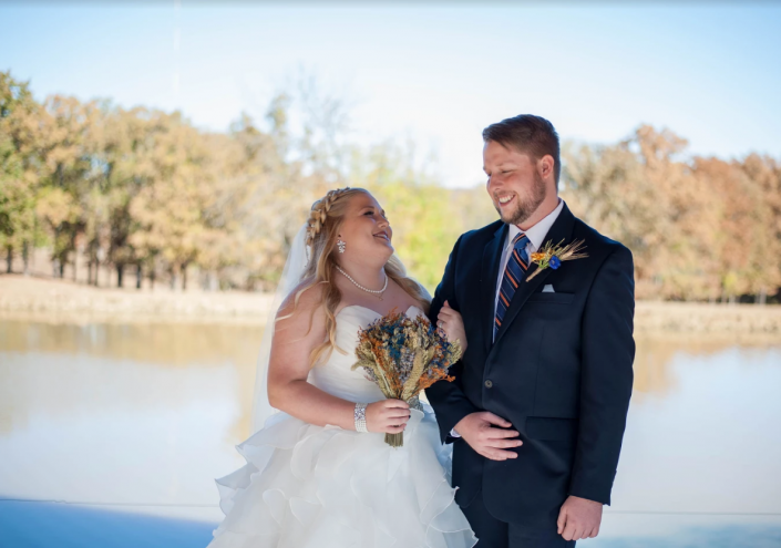 Bride and groom in wedding attire smiling by a lakeside with autumn trees in the background