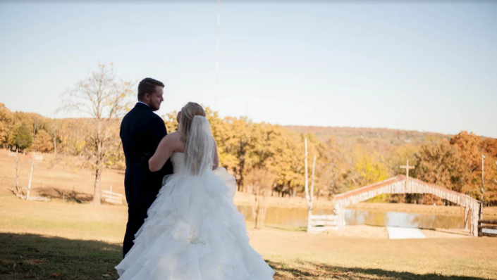 Bride and groom stand outdoors in a sunlit autumn landscape nearby is a pond with a pavilion