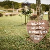 Wooden sign in a grassy outdoor wedding venue reads, “Pick a seat, not a side. You’re loved by both the groom and bride!”