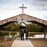 Two people stand at the end of a walkway under a rustic chapel cross by a lakeside.