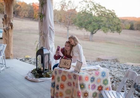 Two people sitting at a decorated outdoor table with lanterns, taking a selfie at a rustic patio overlooking fields