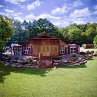 Rustic wooden cabin with red-trimmed windows, stone landscaping, and a green lawn under a blue sky