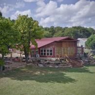Small red-roofed cabin with a porch, stone steps, and a grassy yard under cloudy skies.