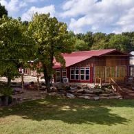 Red-roofed large cabin with large windows in a grassy yard, surrounded by trees under a partly cloudy sky