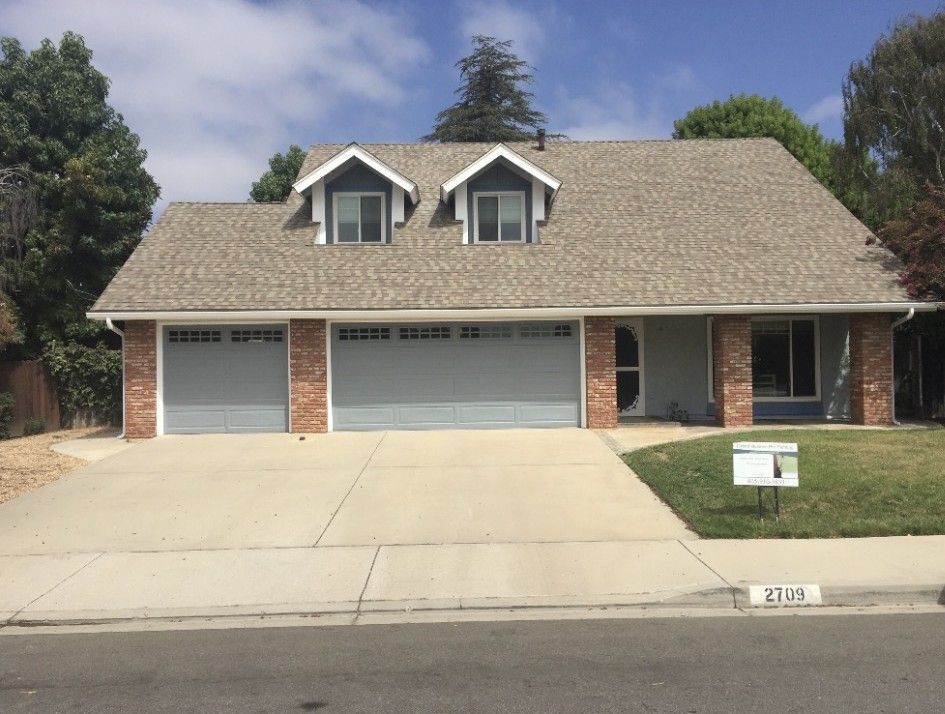 Suburban two-story brick house with a three-car garage, driveway, lawn, and mailbox under a blue sky