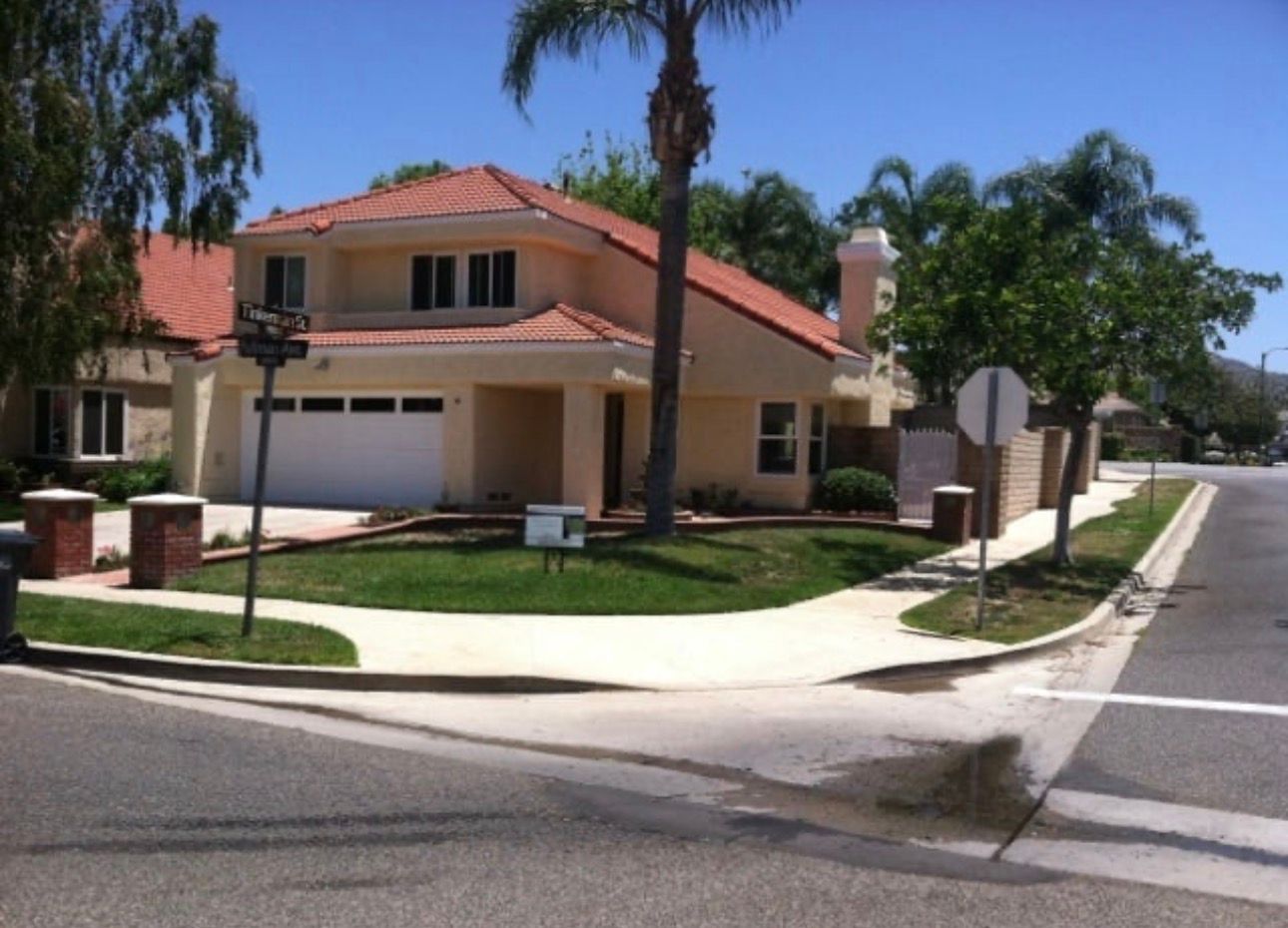 Suburban house with a red-tiled roof, palm trees, and a driveway on a sunny street corner