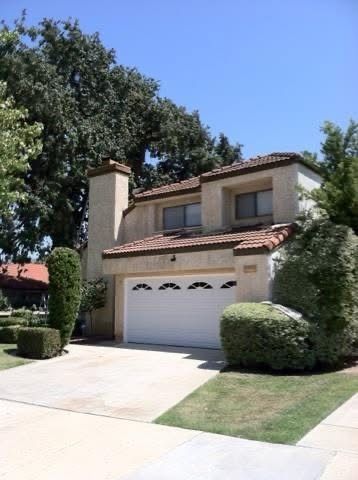 Two-story beige stucco house with a white garage, red-tiled roof, and a driveway in a leafy suburban setting