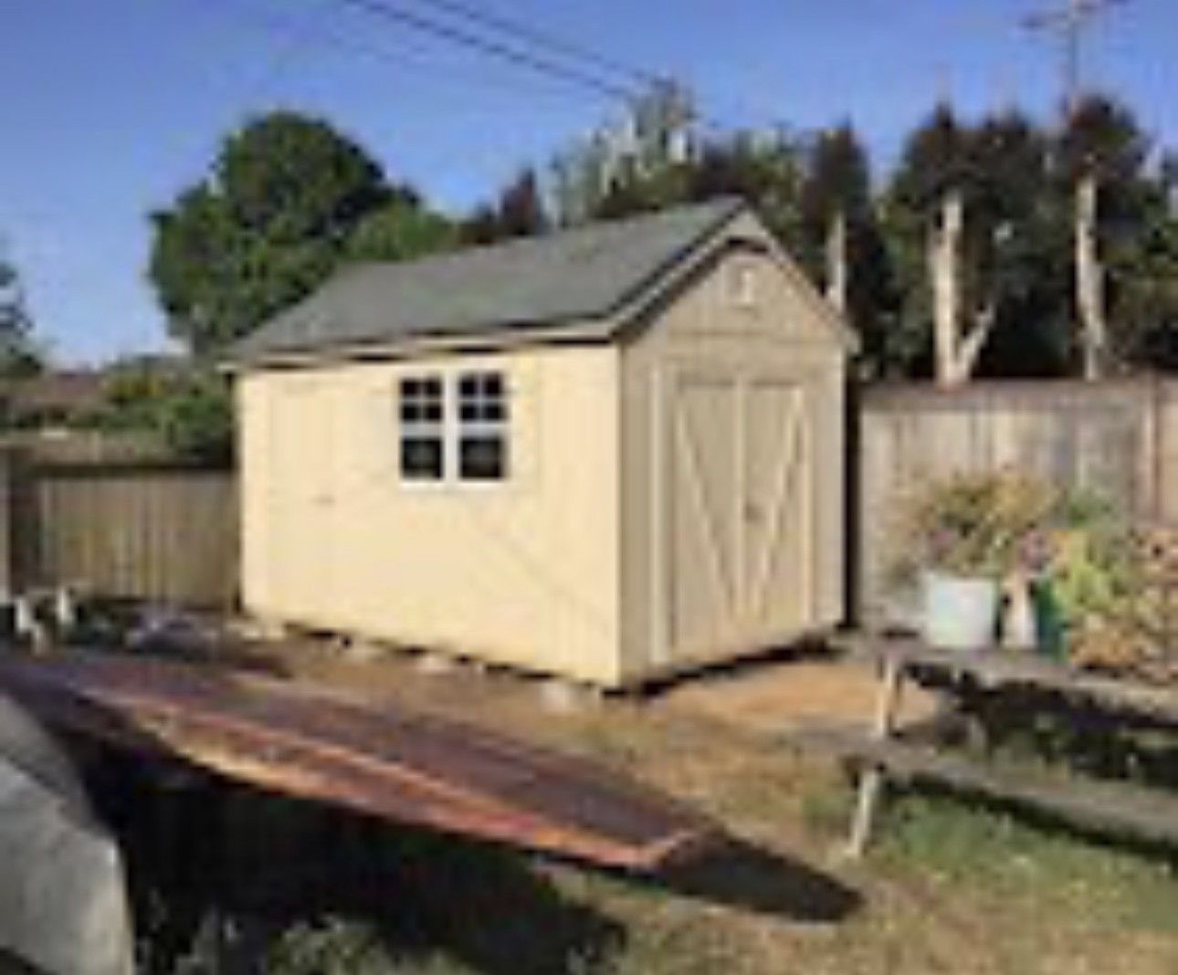 Beige garden shed with white-trimmed windows in a backyard, beside a wooden fence and picnic table