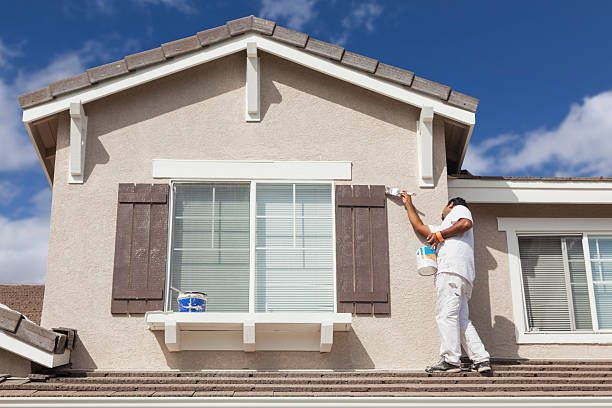 Painter on a rooftop painting a house exterior white under a blue sky.