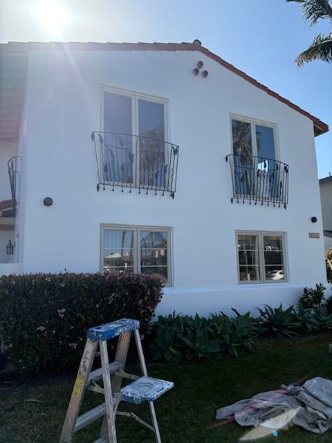 White two-story house with balcony windows, hedge, and a ladder in the front yard under bright sunlight