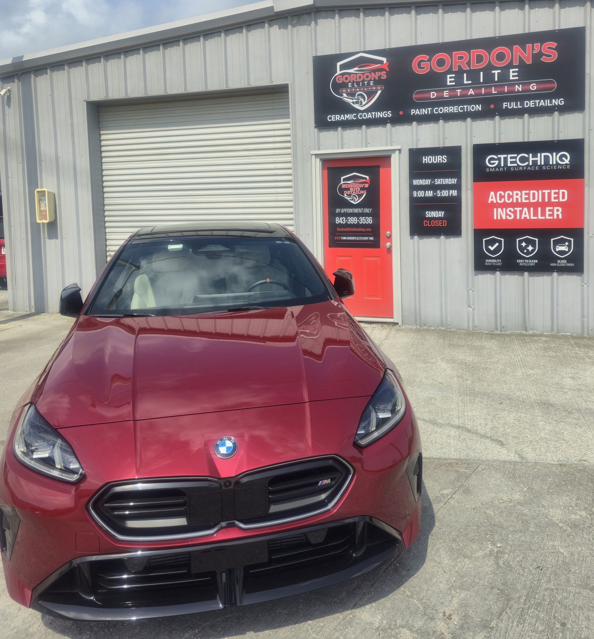 Red BMW sports car parked outside Gordon’s Mufflers shop with red signage and a roll-up door