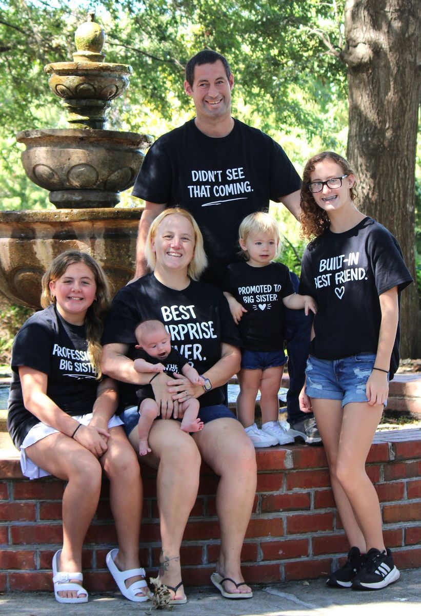Five people in matching black shirts posing on a brick wall by a fountain, outdoors in a park.