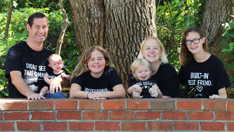 Five people with a baby pose behind a brick wall in a leafy park, smiling at the camera.