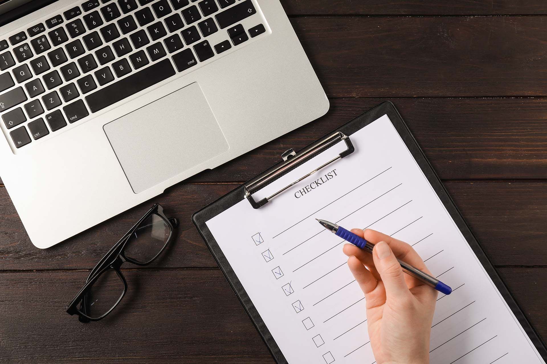 Hand writing on a checklist clipboard beside a laptop and glasses on a dark desk