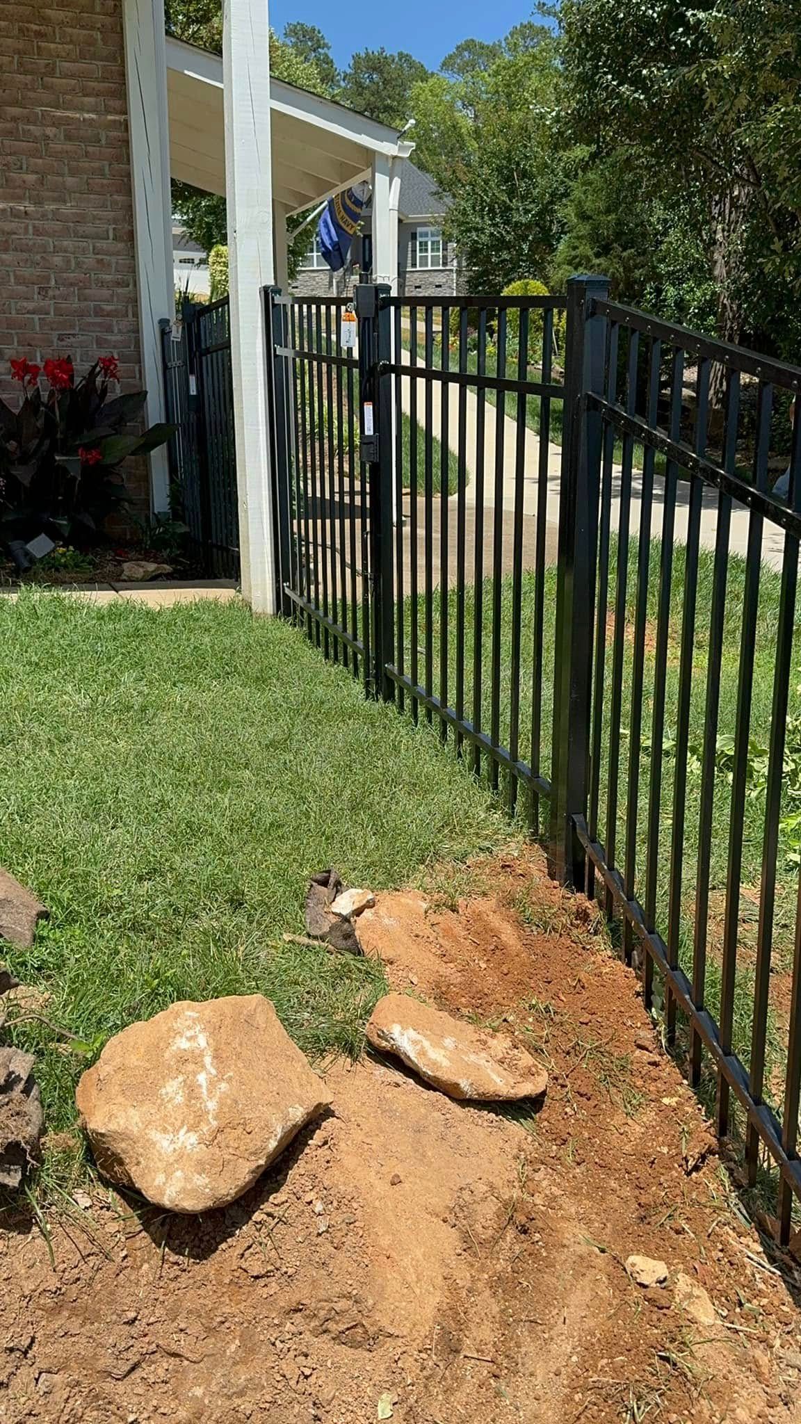 Partially dug yard beside a black metal fence, with soil and rocks next to a grassy lawn and house porch.