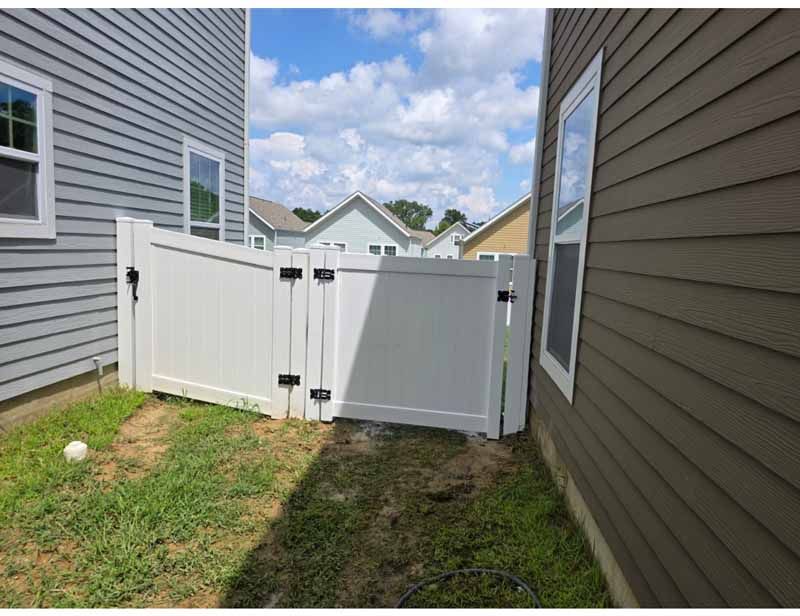 White vinyl gate between two houses in a narrow grassy side yard under a cloudy sky