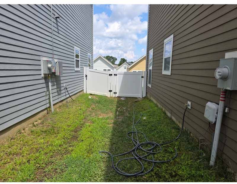Narrow grassy alley between two houses with utility meters, hoses, and a white gate at the end