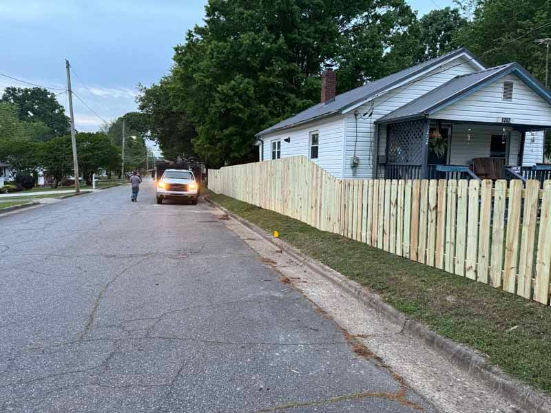 Quiet residential street with a white house, wooden fence, and car driving away at dusk