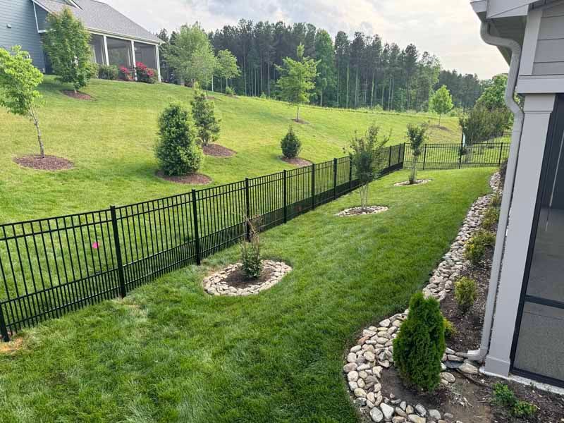 Backyard with black fence, green lawn, small trees, and stone-edged planting beds beside a house