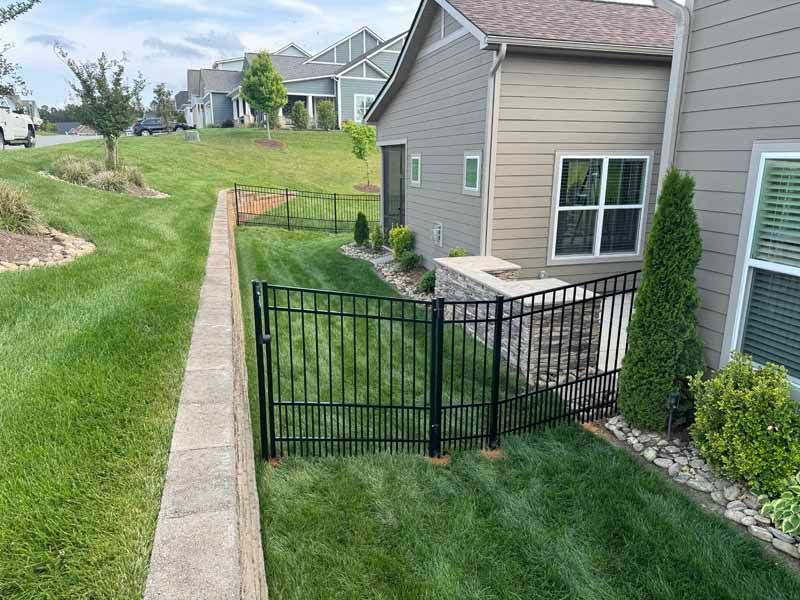 Black metal gate beside a tan townhouse, opening onto a grassy side yard and concrete sidewalk.