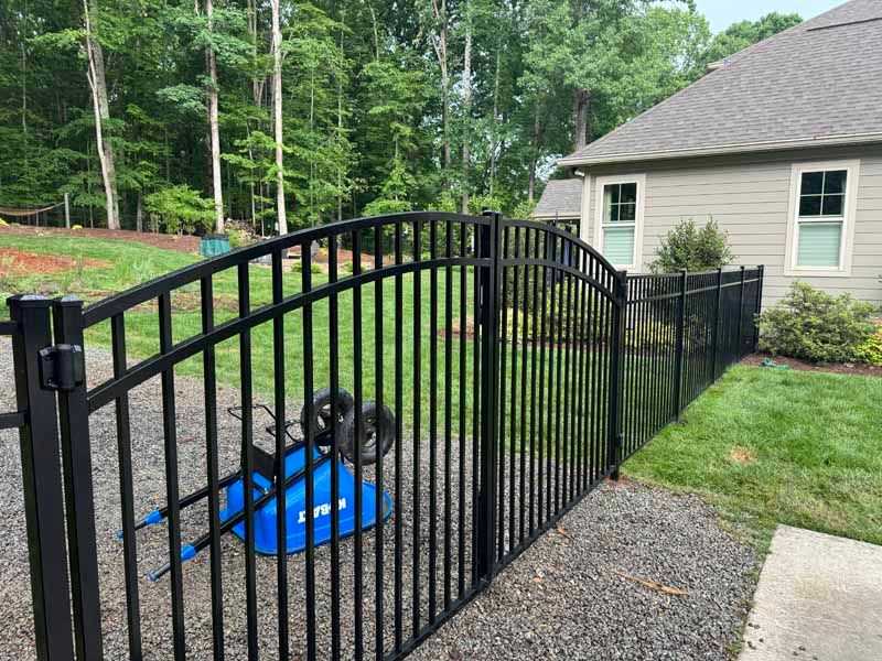 Black metal gate in a gravel yard beside a house, with a blue tarp-covered object behind it.