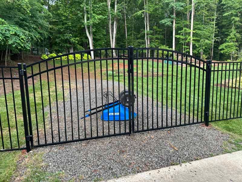 Black arched metal gate across a gravel driveway, with a blue object behind it and trees in the background