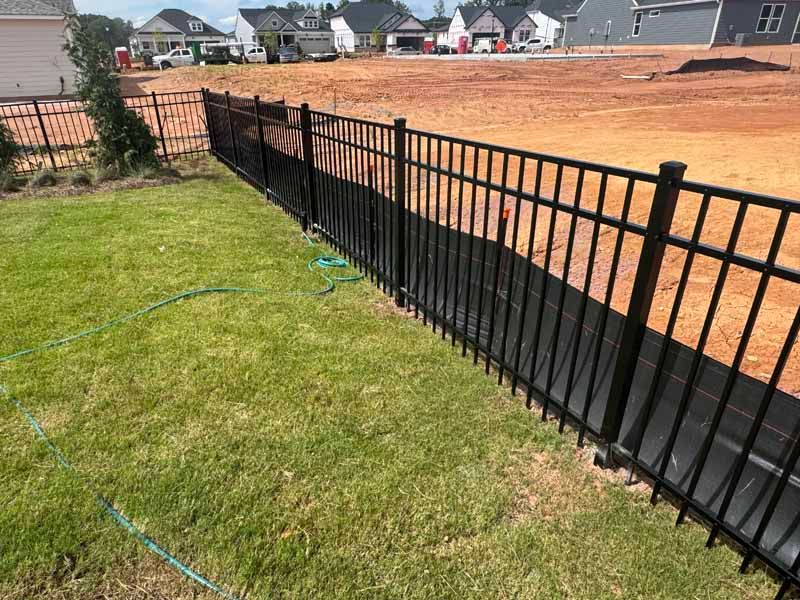 Black metal fence along a grassy yard beside a large dirt construction site in a suburban neighborhood.