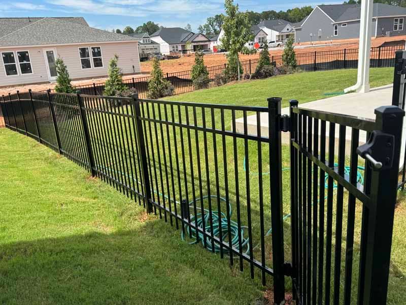 Black metal fence along a sloped green lawn beside houses and a wooden privacy fence.