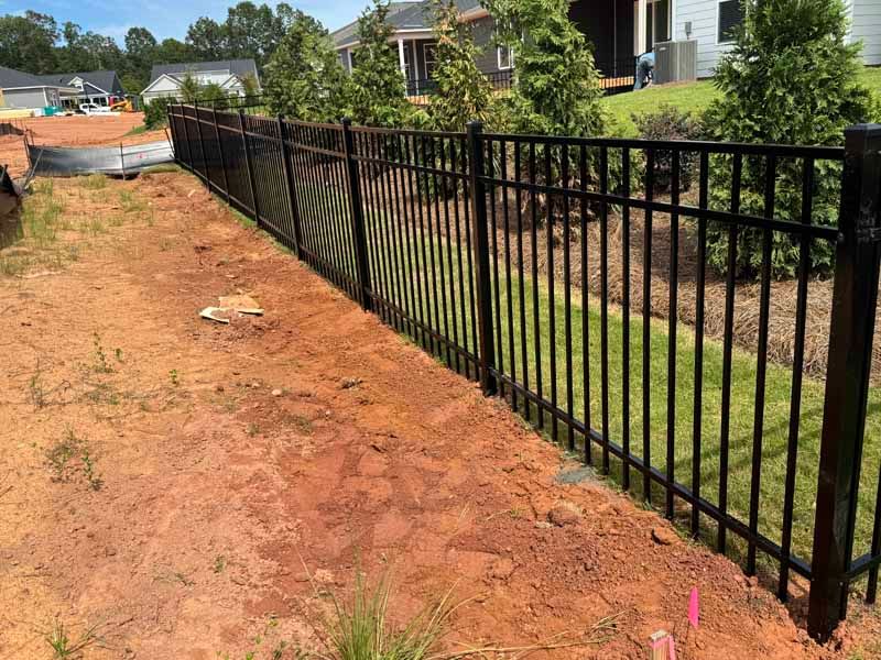 Black metal fence along a dirt path beside a grassy residential yard and houses
