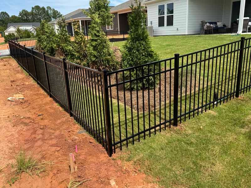 Black metal fence along a grassy yard beside a red dirt path in a suburban neighborhood