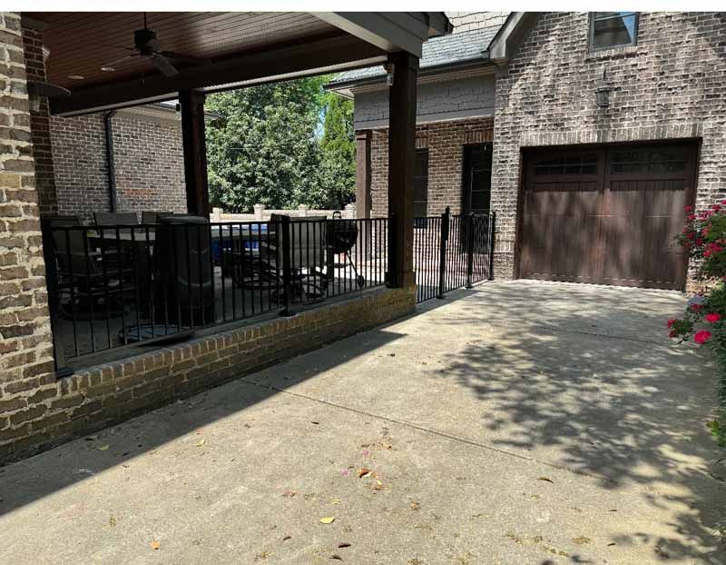 Brick driveway beside a covered patio and garage with black metal railing in a sunny yard