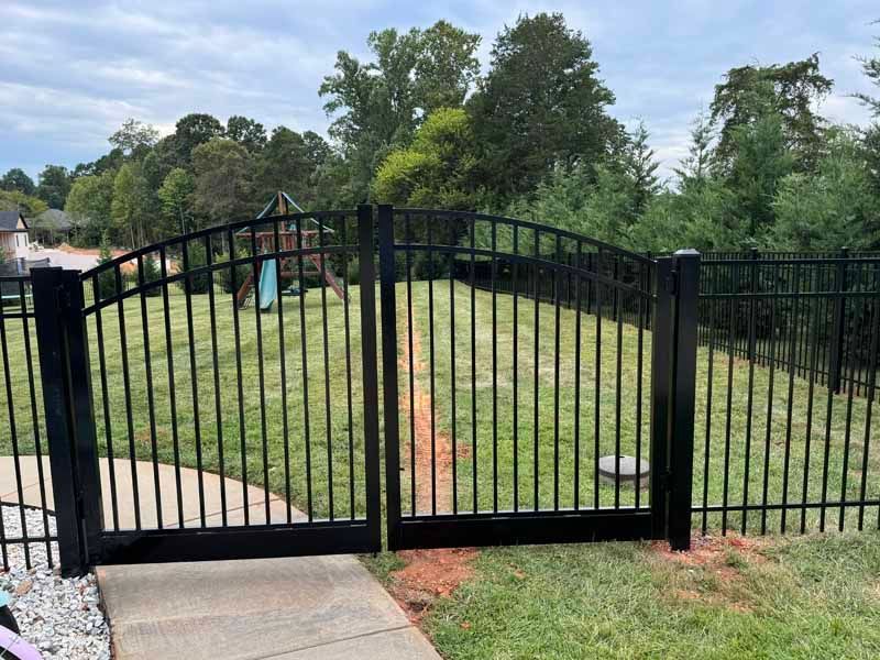 Black metal gate at the end of a concrete path, opening to a grassy yard with trees beyond.
