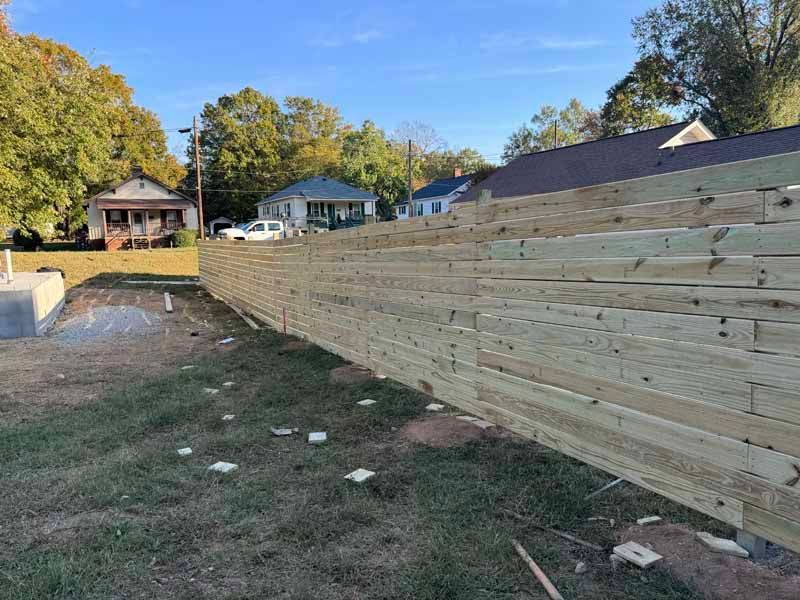 Long wooden retaining wall along a grassy yard under a blue sky, with houses and trees in the background