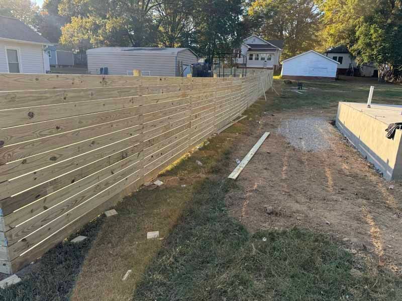Wooden privacy fence under construction beside a dirt yard, with posts and boards laid out in a residential area.