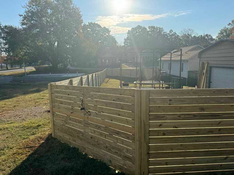 Sunlit backyard with a wooden privacy fence and sheds, casting long shadows across the grass.