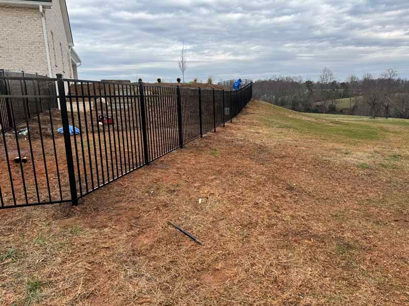 Black metal fence beside a beige house on a sloping, brown grassy yard under an overcast sky