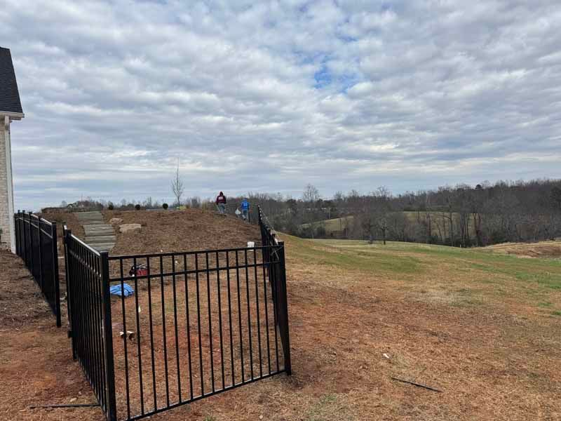 Black metal fence opens onto a grassy yard under a cloudy sky, with a house at left and trees in the distance