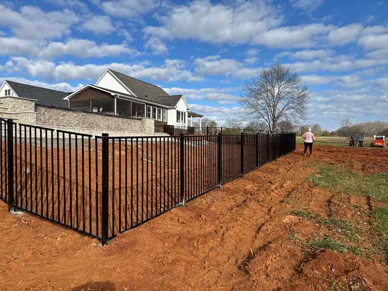 Black metal fence beside a house along a dirt path, with one person walking in the distance.
