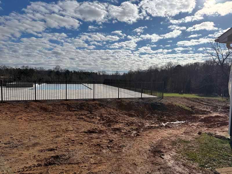 Bare dirt lot beside a black fence under a cloudy blue sky with trees in the distance