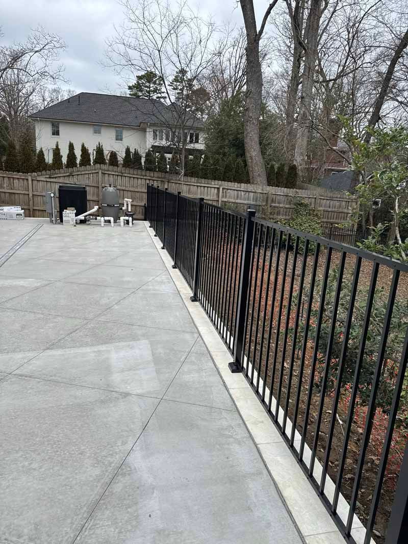 Stone patio with black railing overlooking a wooded yard and house in the distance