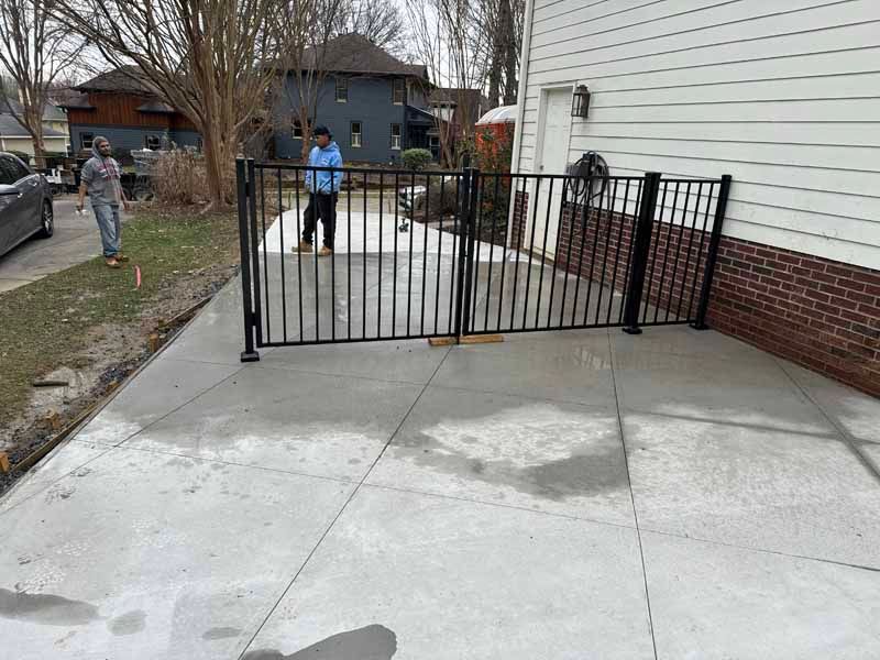 Black metal gate blocking a concrete driveway beside a white house, with two people standing behind it.