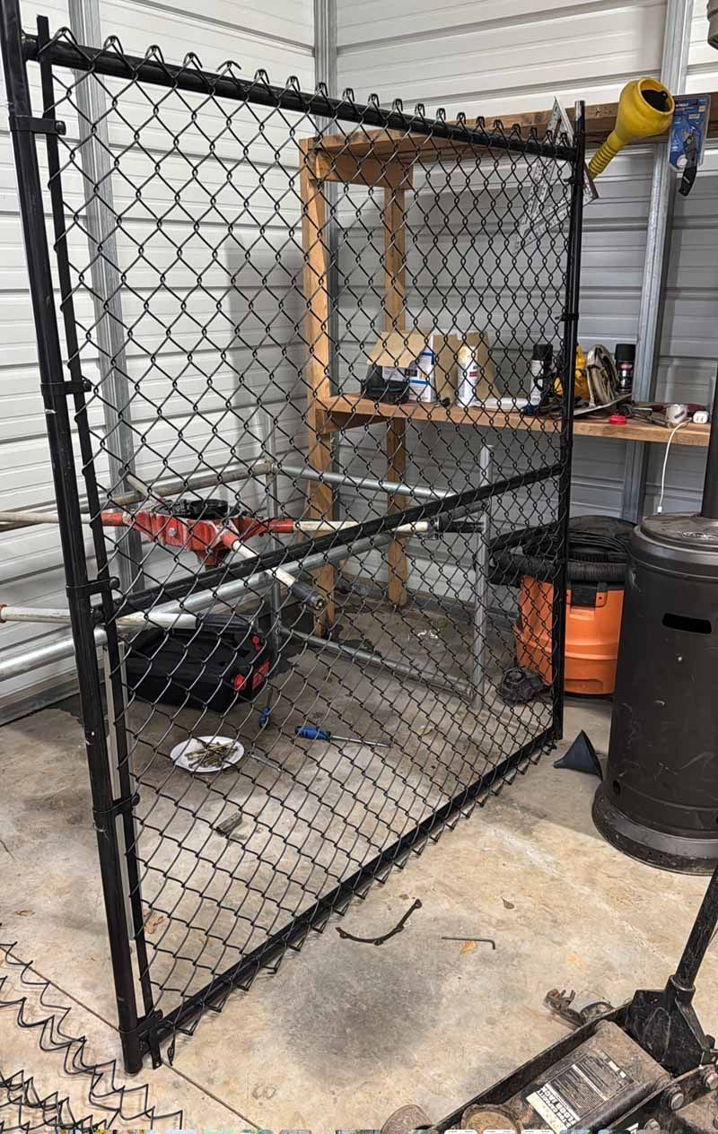 Black chain-link fence panel leaning in a workshop/garage with shelves and tools behind it