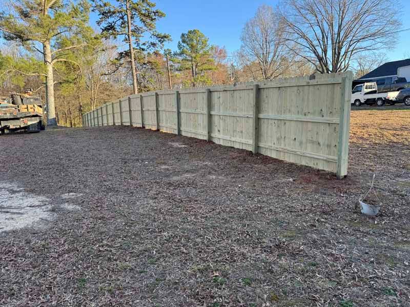 Long beige wooden fence along a gravel lot with trees and parked vehicles in the background.