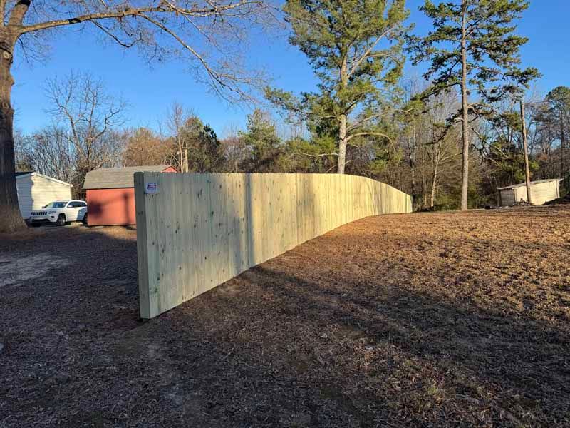 Curved wooden privacy fence in a yard, with trees and blue sky in the background.