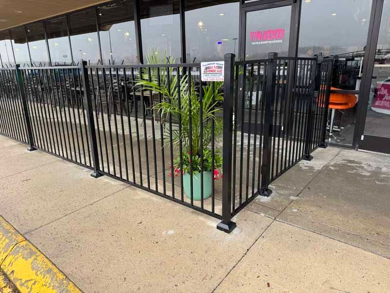 Black metal fenced patio outside a storefront, with potted plants and outdoor seating by the entrance.