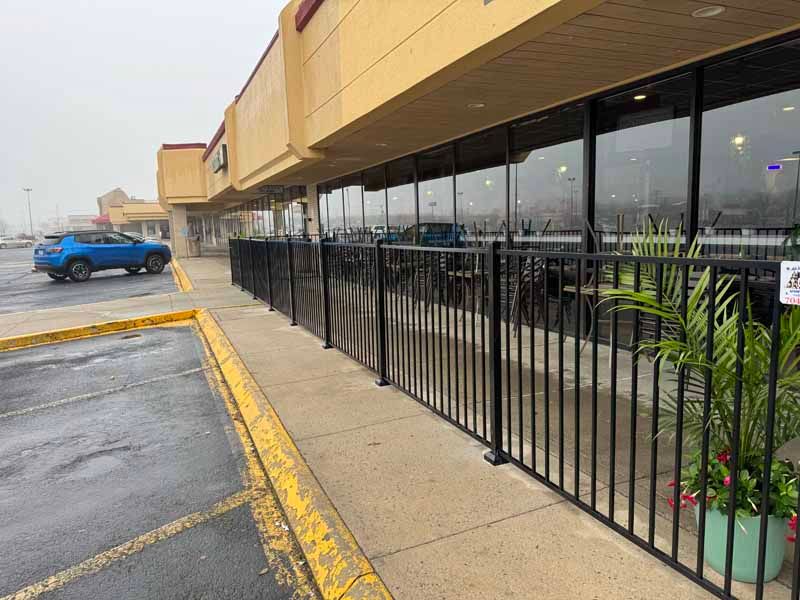 Strip mall storefronts with black railings, wet pavement, and a blue car in the parking lot.