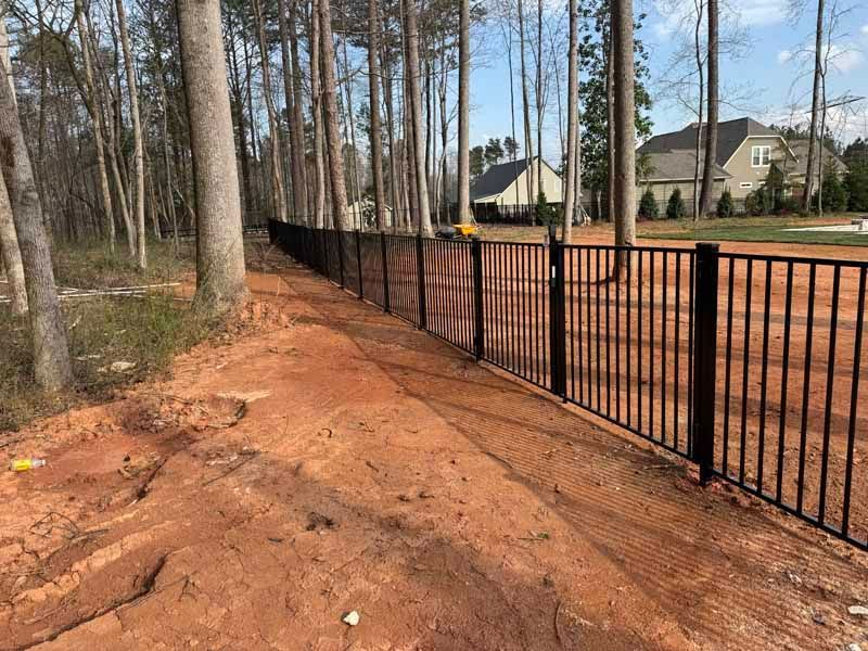 Dirt path beside black metal fence along wooded suburban yard under clear blue sky