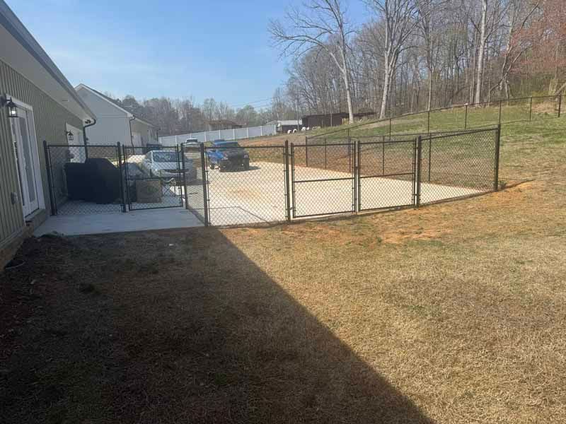 Backyard with concrete patio, black chain-link fence, house siding, parked car, and grassy lawn under sunny sky
