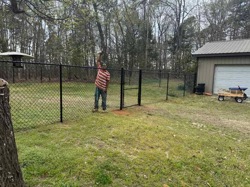 Person in striped shirt opening a chain-link gate in a grassy yard beside a garage.