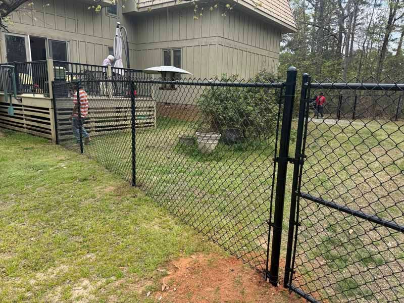 Chain-link fence with open gate beside a house and grassy yard; patio and umbrella in the background
